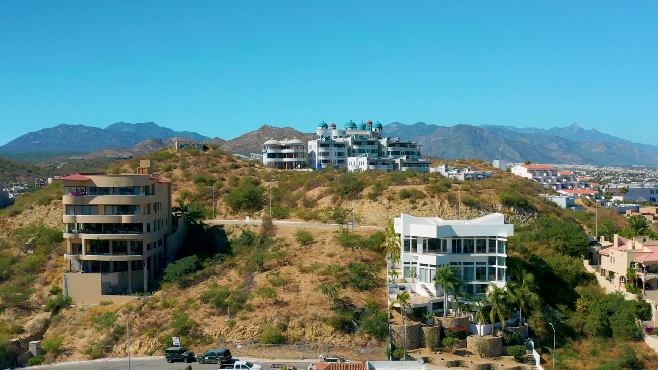 Opening view of San Jose del Cabo hillside homes with a road at the bottom edge, dry terrain, distant mountains, clear blue sky, and no visible people.
