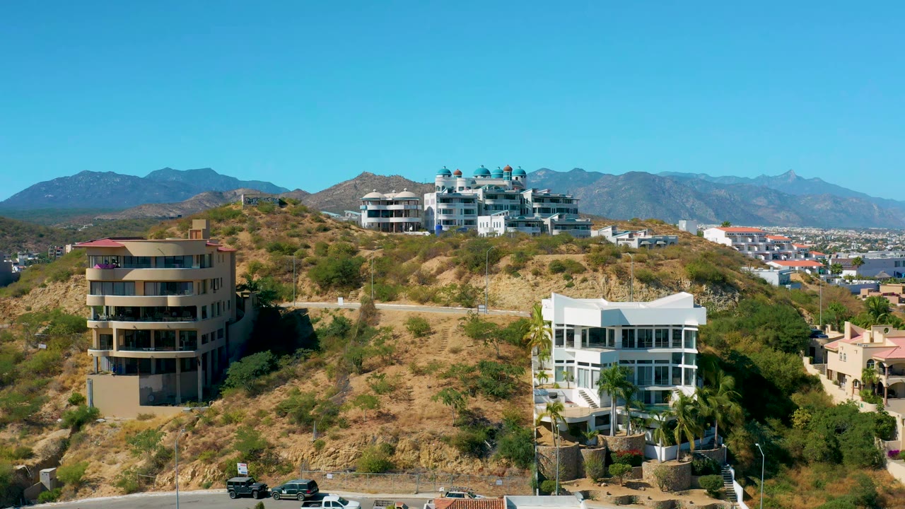 San Jose del Cabo hillside residences fill the middle of the frame, with dry brush slopes, a few vehicles near the road, mountains in the distance, and no visible people.