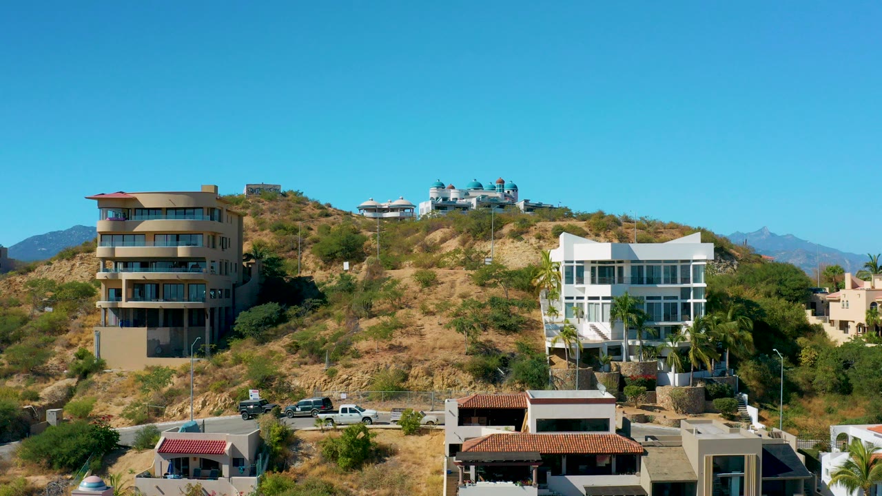 Wide elevated view of hillside residences in San Jose Del Cabo, with tan and white buildings, dry vegetation, foreground roofs, distant mountains, and no visible people.