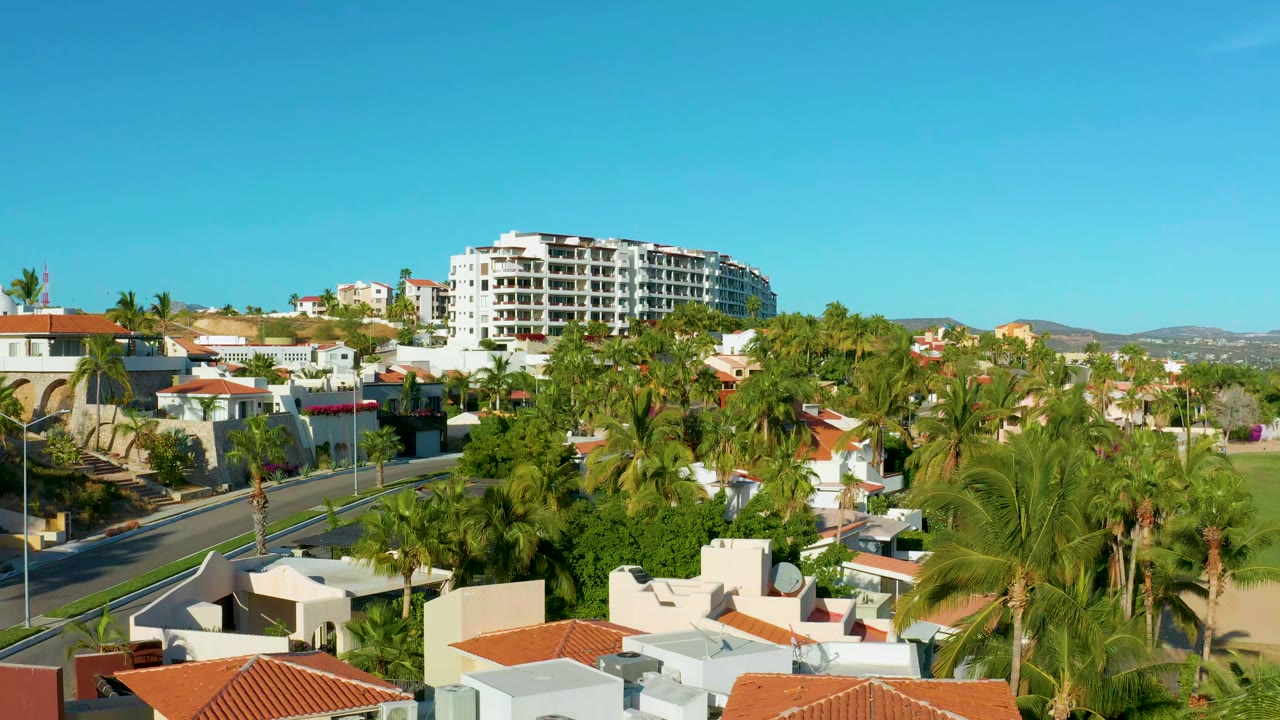 Opening aerial view over San Jose del Cabo homes with palm trees, a curving road at left, a large white building in the background, and no visible people.
