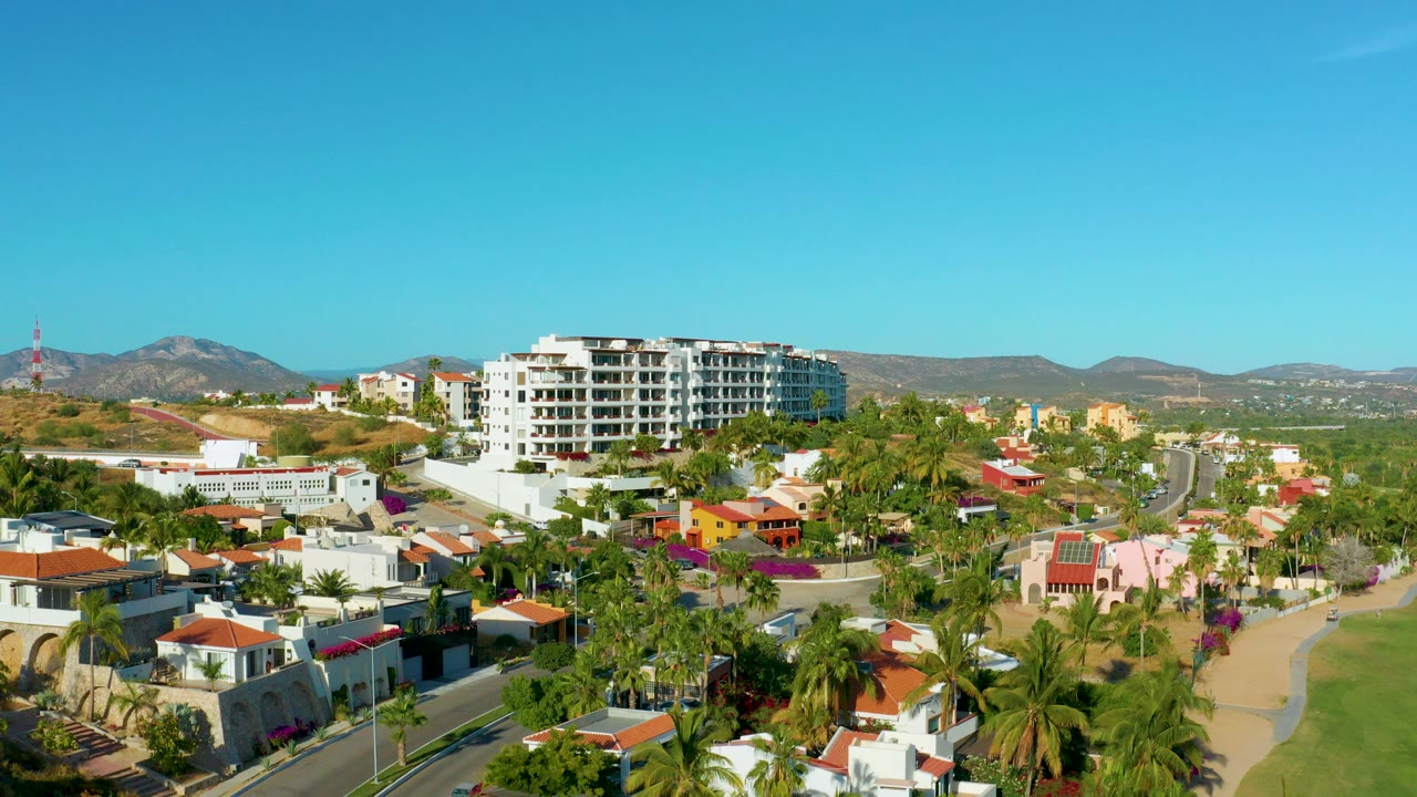 Opening aerial frame over San Jose del Cabo homes, palm trees, red tile roofs, a white mid-rise building, dry hills, and clear blue sky with no visible people.