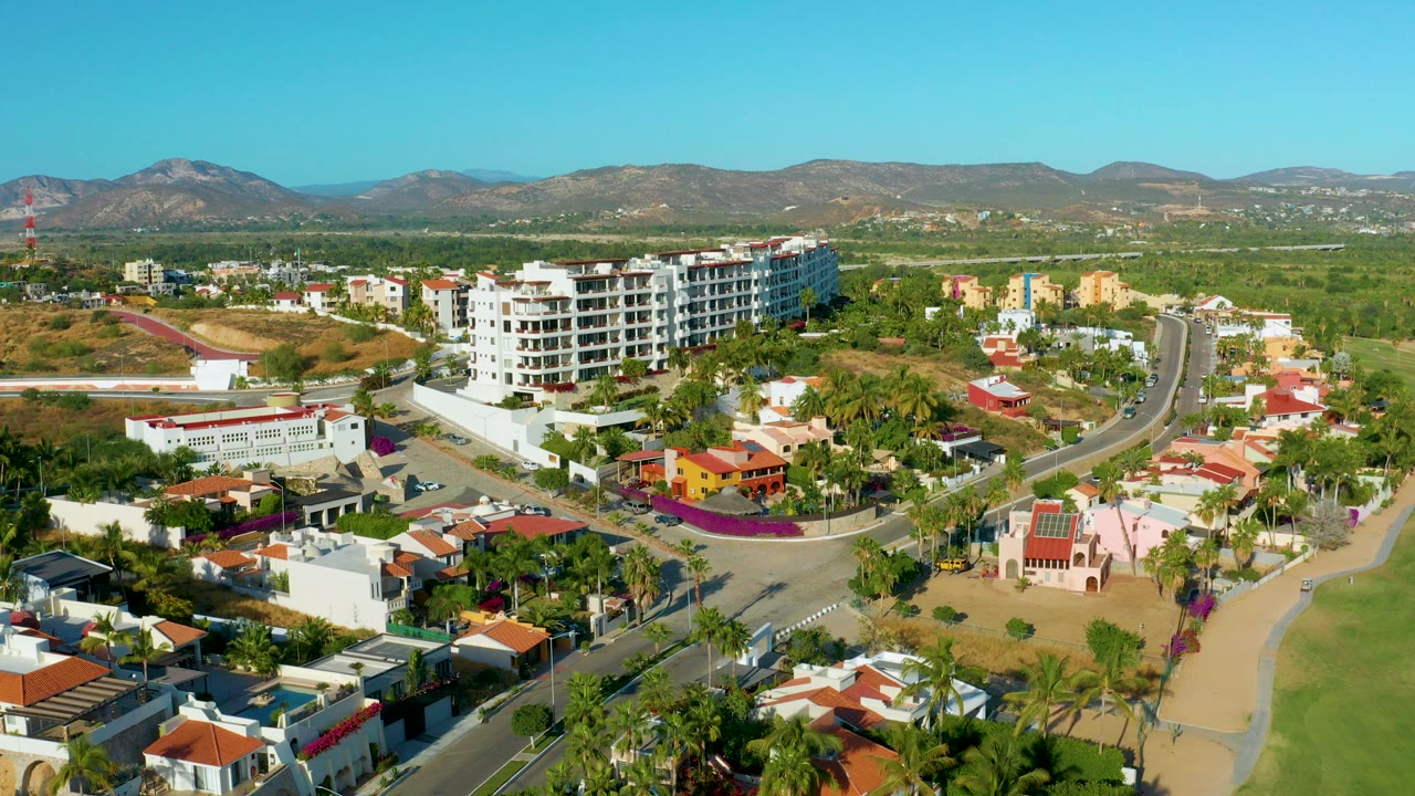 Opening aerial frame over San Jose Del Cabo homes, a curved road, palm trees, a white multi-story building, distant mountains, and no visible people.