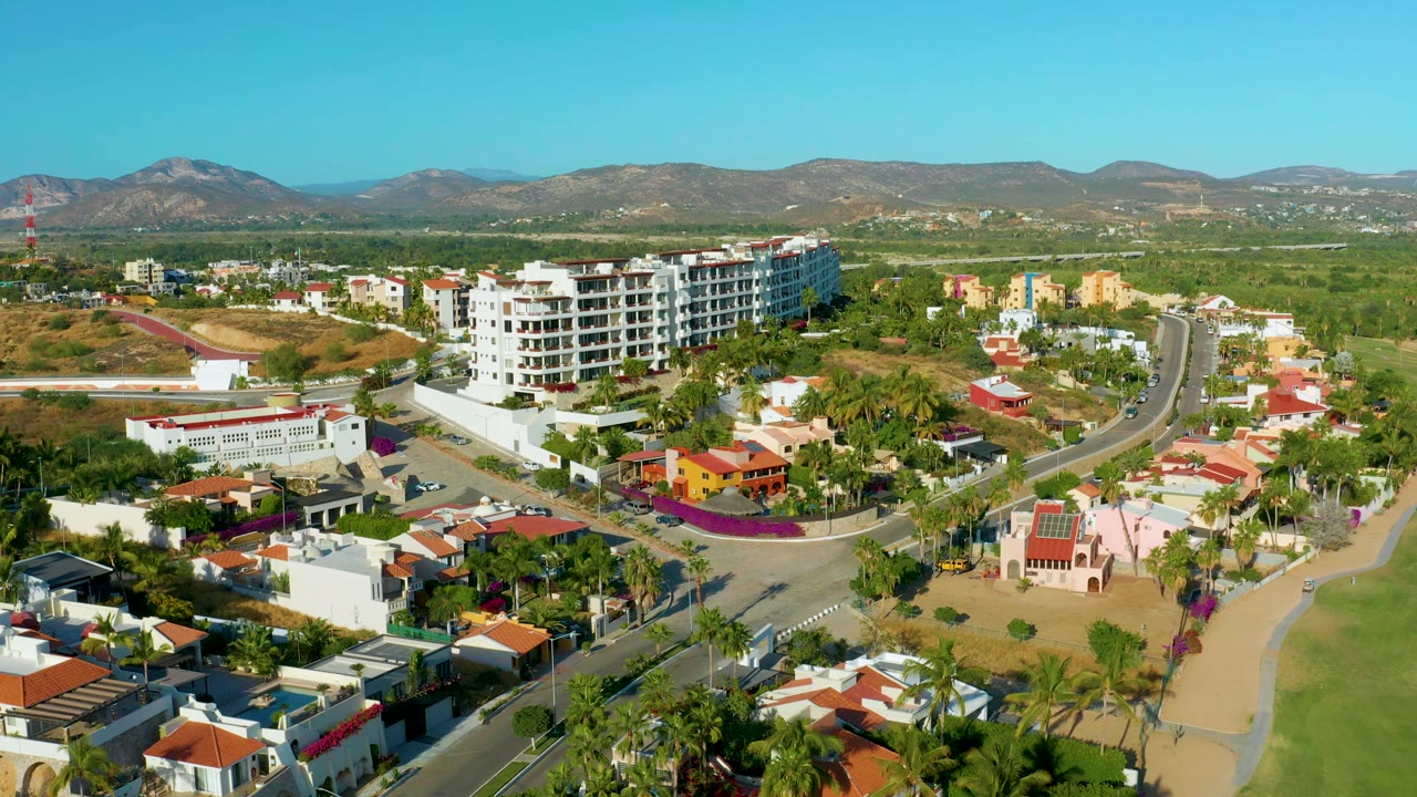 Aerial view in San Jose Del Cabo with the white multi-story building near center, small cars on the curved road, palms, sandy lots, mountains, and no visible people.