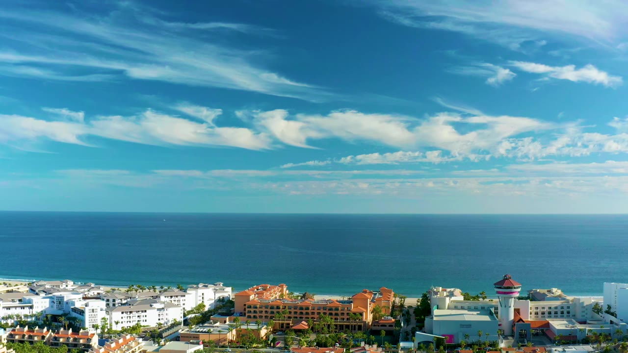 San Jose Del Cabo rooftops and greenery sit along the lower frame while the ocean and streaked white clouds fill most of the view, no visible people.