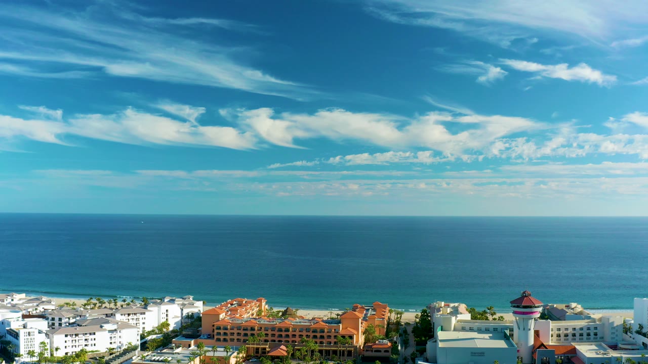 Mid-clip San Jose Del Cabo view with calm blue sea, a level horizon, low white and orange coastal buildings near the bottom, and no visible people.