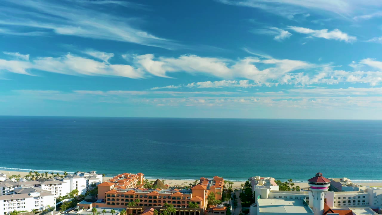 San Jose del Cabo beach view with the ocean filling most of the frame, white clouds stretched across the sky, and resort-style buildings along the lower edge, with no visible people.