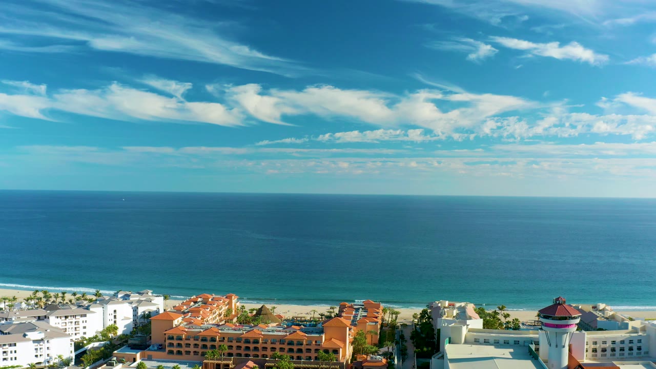 Mid-clip view over San Jose del Cabo with calm blue water, a thin white surf line, coastal buildings at the bottom edge, and high wispy clouds, with no visible people.