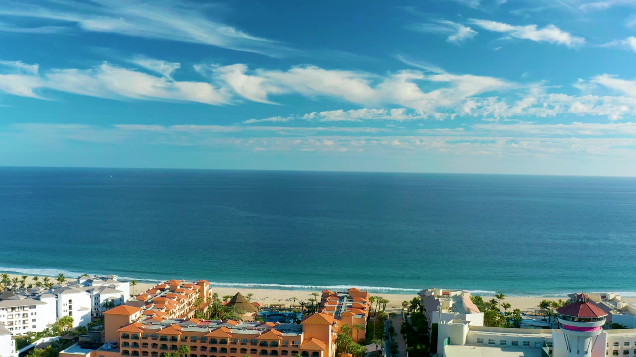 San Jose Del Cabo shoreline view with a broad blue sea horizon, thin surf line, orange-red roofs, white beachfront buildings, scattered palms, and no visible people.