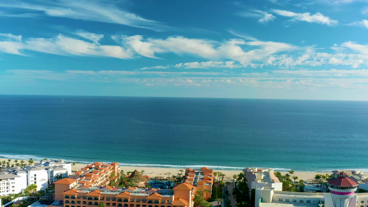 Aerial San Jose Del Cabo frame with calm ocean water, a pale beach strip, red-roofed coastal property, bright white buildings, palm trees, and no visible people.