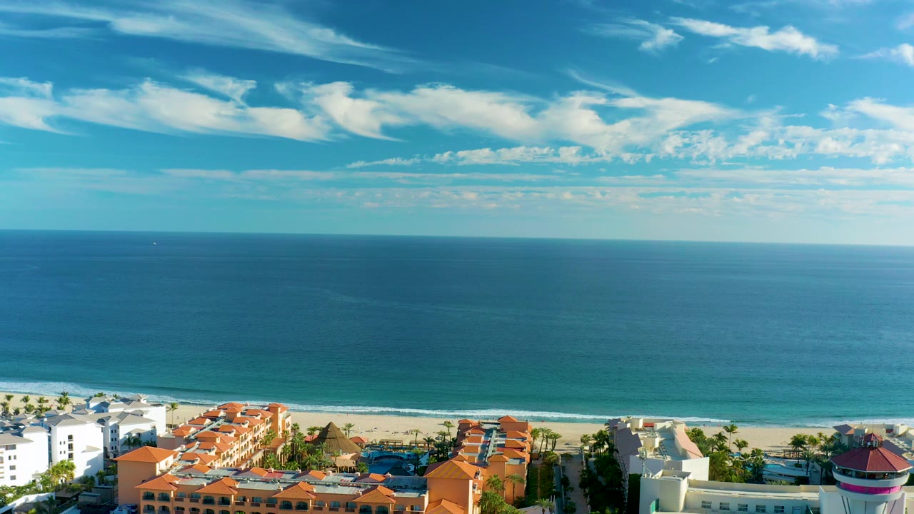 Mid-clip aerial view in San Jose Del Cabo with white and red-roofed coastal buildings below a wide blue ocean, soft cloud bands, and no visible people.