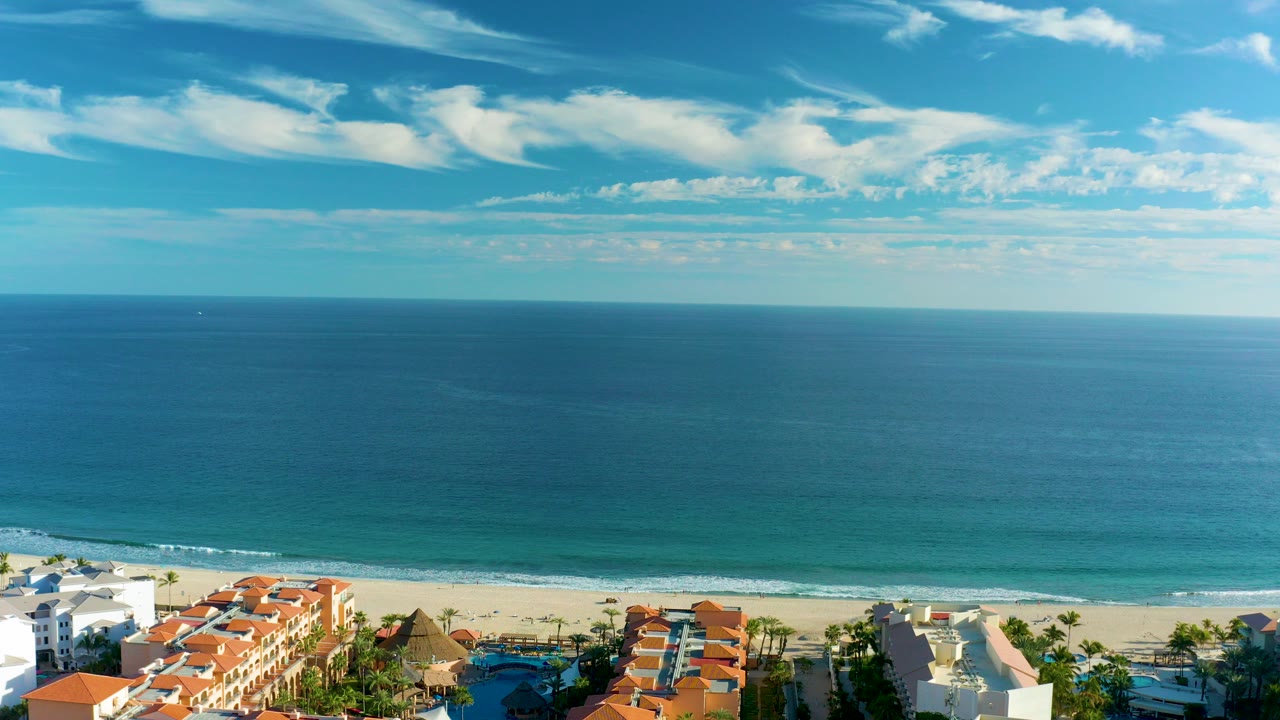 Aerial frame over San Jose del Cabo showing blue ocean, sandy shore, red-tiled resort roofs, white buildings, palm trees, scattered clouds, and no visible people.