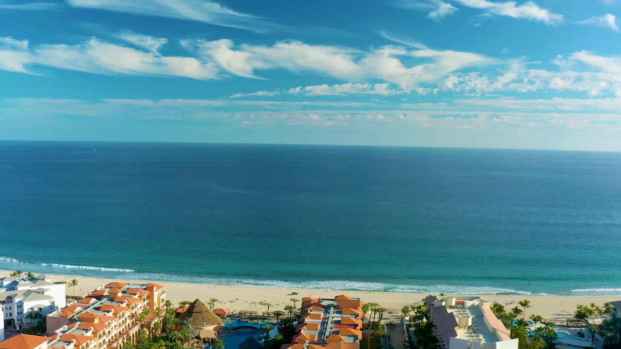 San Jose del Cabo coastline frame with a level ocean horizon, thin surf line, red roof resort structures, white facades, palm trees, and no visible people.
