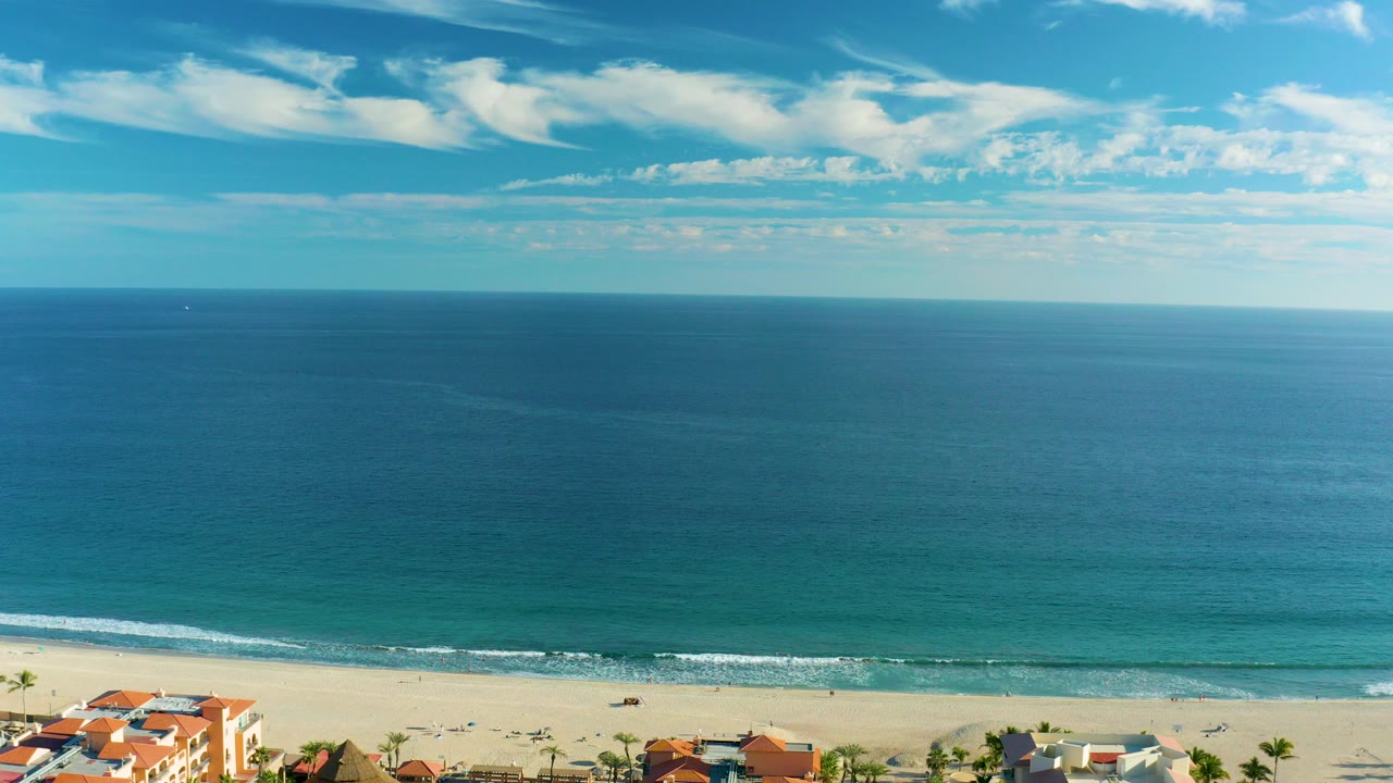 San Jose del Cabo coastline seen from an elevated view, with red-roof buildings along the bottom, a pale beach, blue ocean, scattered clouds, and no visible people.