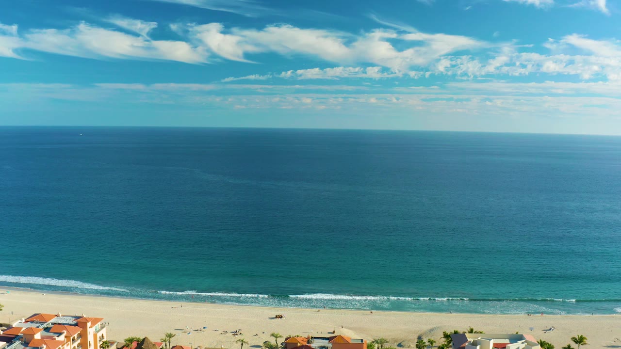 Elevated San Jose del Cabo coastal frame with a small cluster of red-roof buildings at the lower edge, a bright sand strip, blue-green water, and no visible people.