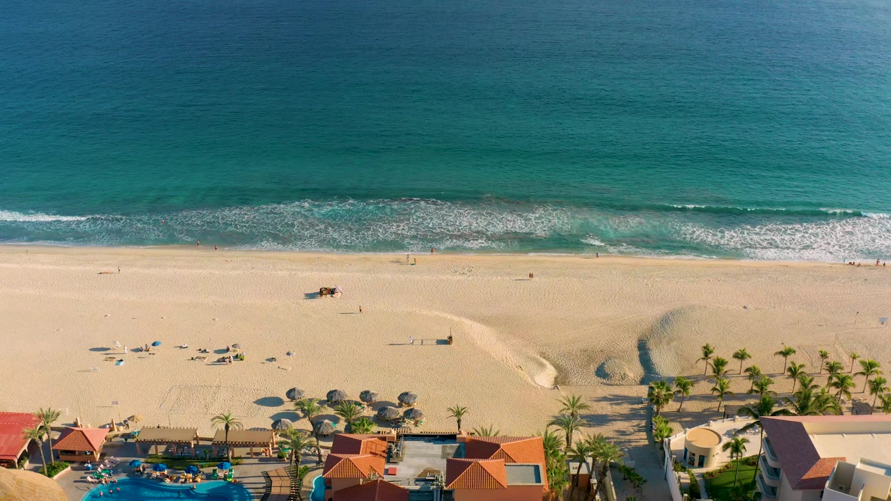 High drone view in San Jose Del Cabo with turquoise ocean across the upper frame, foamy surf at the beach edge, broad sand, clustered umbrellas, palms, and resort rooftops.