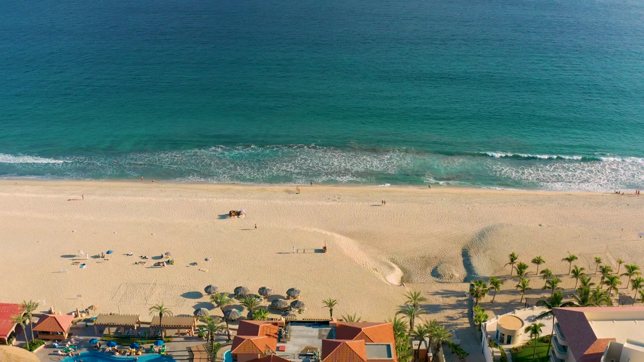 Drone view of waves rolling toward an open beach with palm trees and resort structures at the bottom.