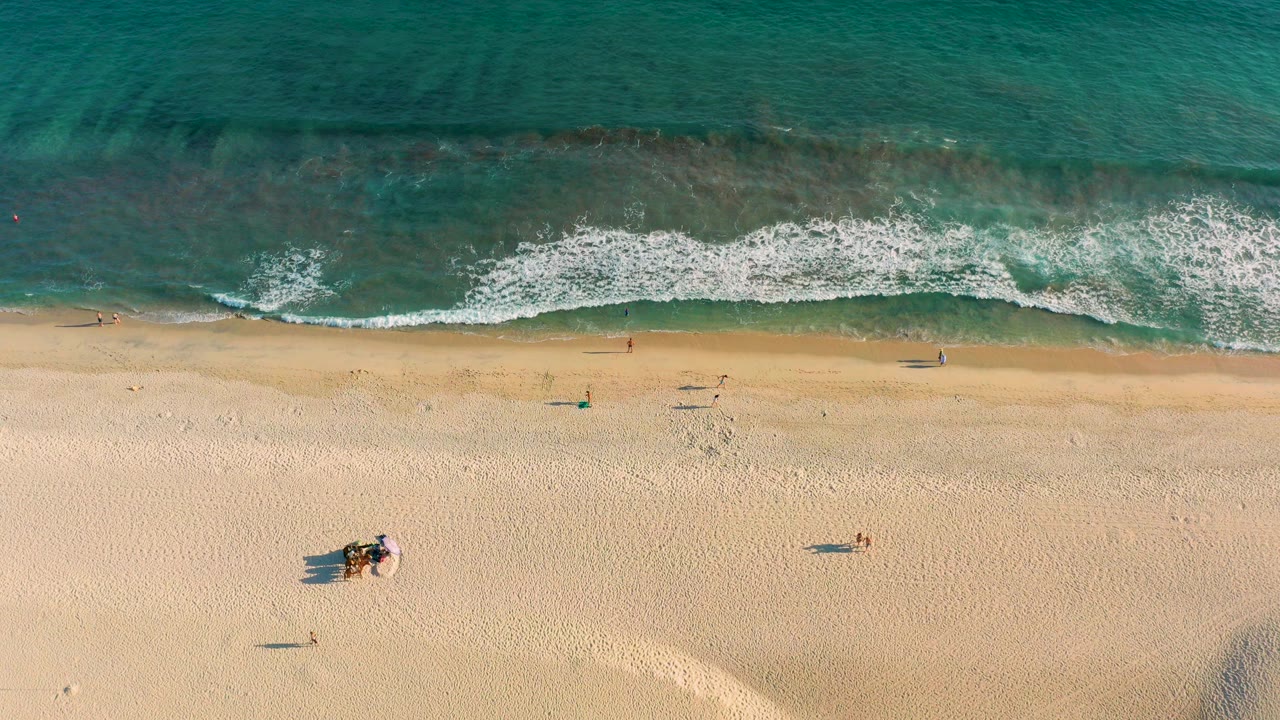 Overhead view of a San Jose del Cabo beach at 0:00, with turquoise water and white foam above pale sand where a few small beachgoers are visible.