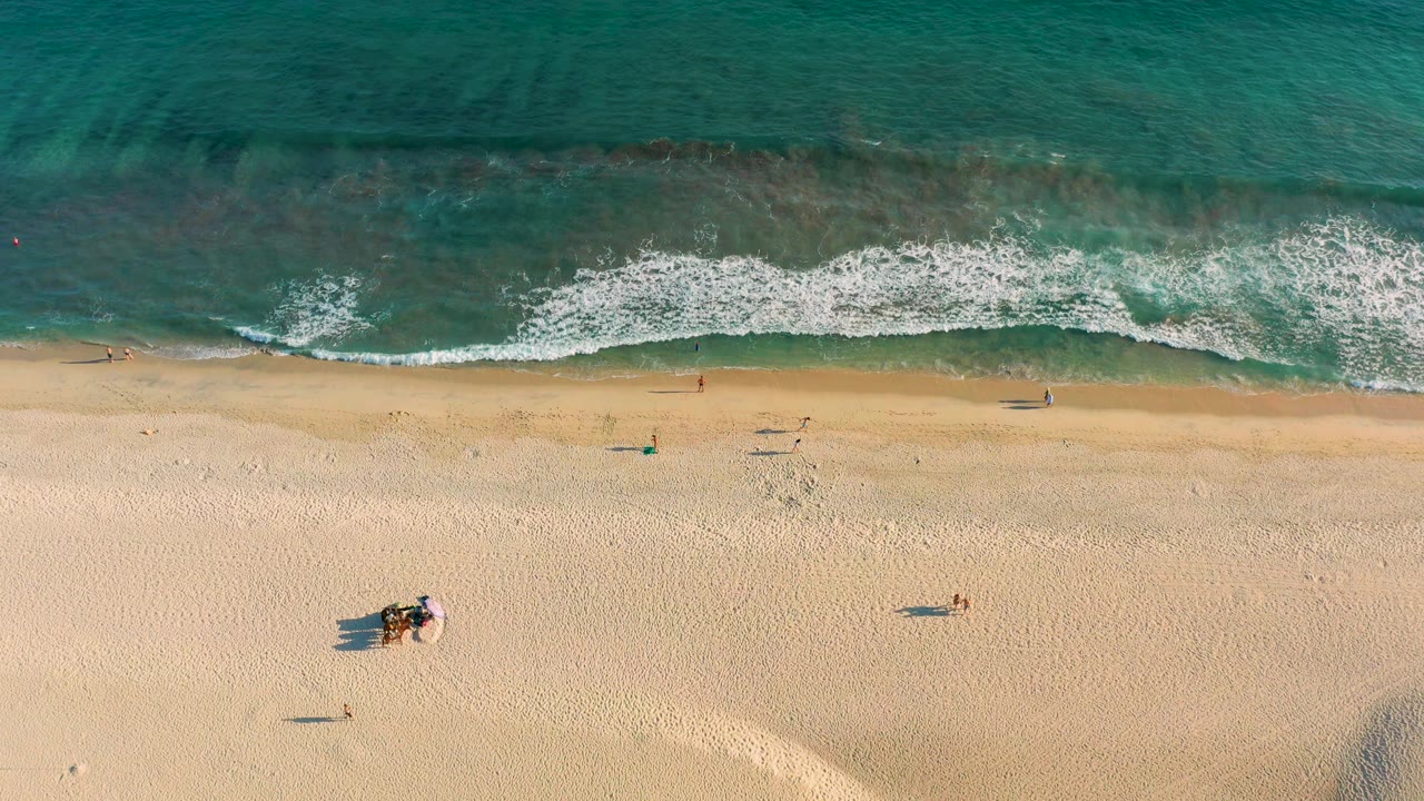 Aerial shoreline with breaking white waves and scattered people on sand.