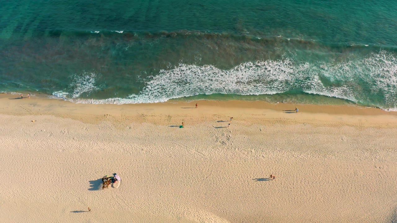 Top-down San Jose del Cabo beach frame at 0:02, with foamy waves across the upper edge and a small cluster of beachgoers lower in the sand.
