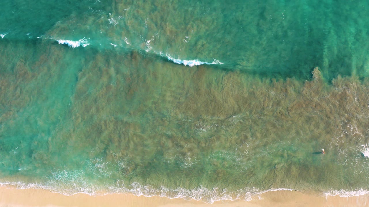 Overhead turquoise ocean with a thin foam line approaching sand.