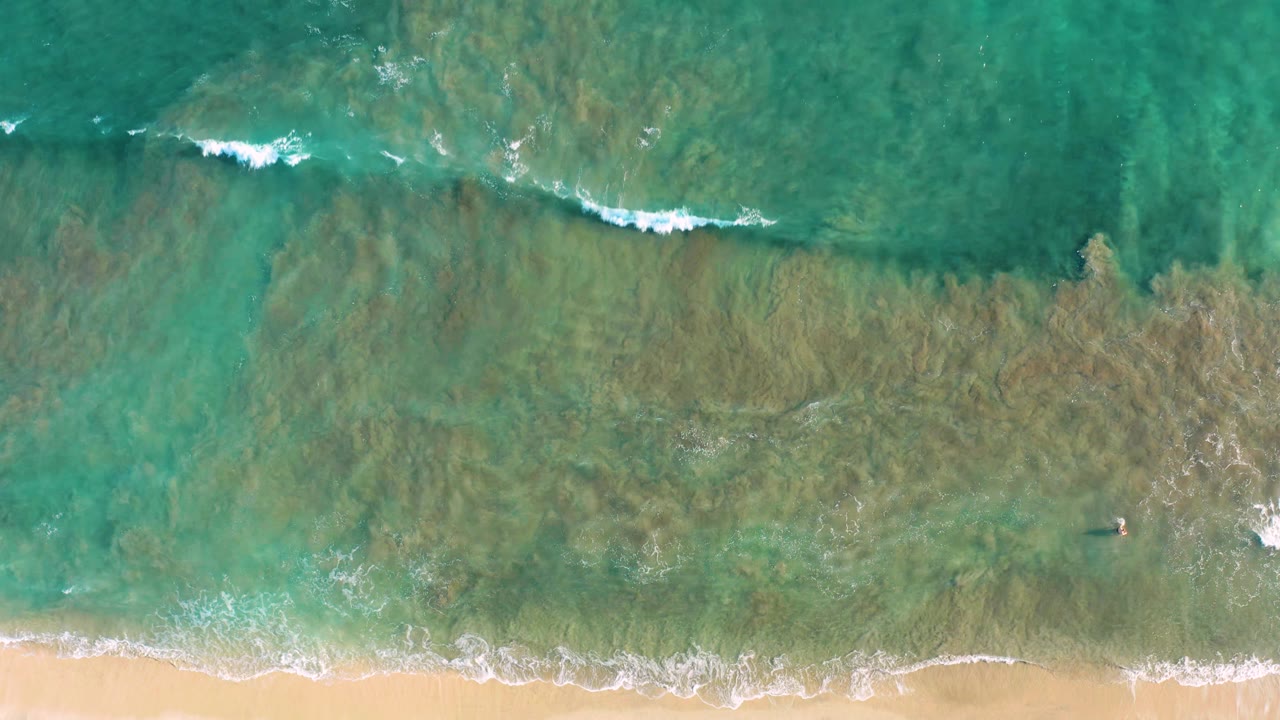 Aerial frame from San Jose del Cabo with green-blue water, a low strip of white surf, submerged darker texture, sandy beach along the lower edge, and no visible people.