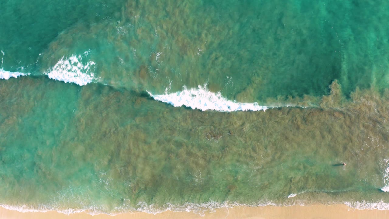 Overhead San Jose del Cabo beach frame with a wider white foam band crossing shallow turquoise-green water, darker reef-like patches underneath, sand below, and no visible people.