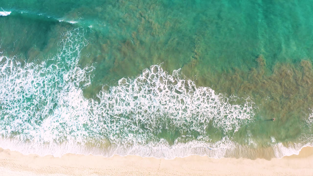 Aerial frame from San Jose del Cabo with a wider white foam band crossing shallow turquoise water above the sandy shore, with no visible people.