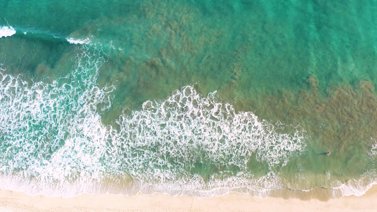 Top-down San Jose del Cabo shoreline view where broken white surf sits between clear green-blue water and a narrow sandy beach, with no visible people.