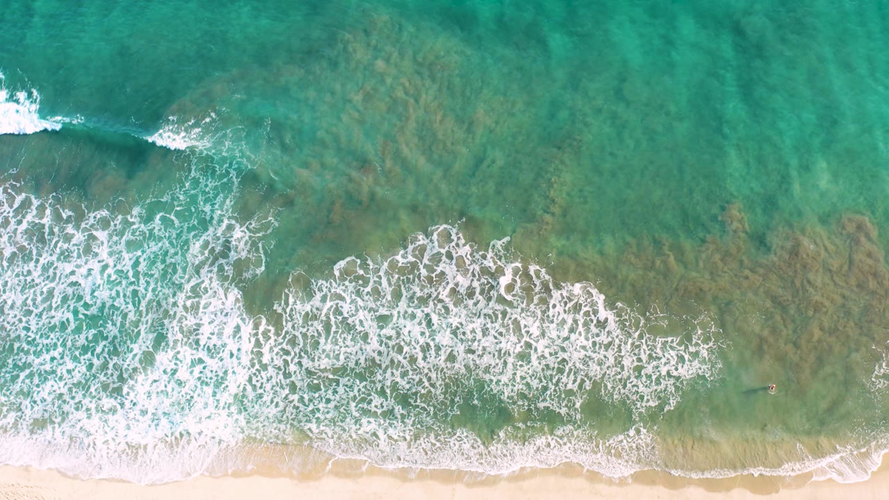 Overhead San Jose del Cabo beach frame with a broad foamy wave patch moving over shallow turquoise water toward pale sand, with no visible people.