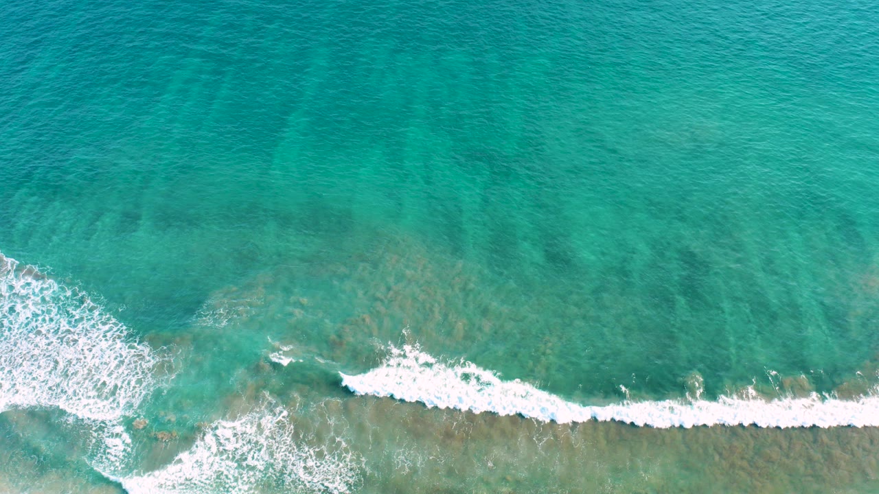 Overhead view of turquoise ocean in San Jose del Cabo with a white wave band breaking across shallow seabed colors and no visible people.