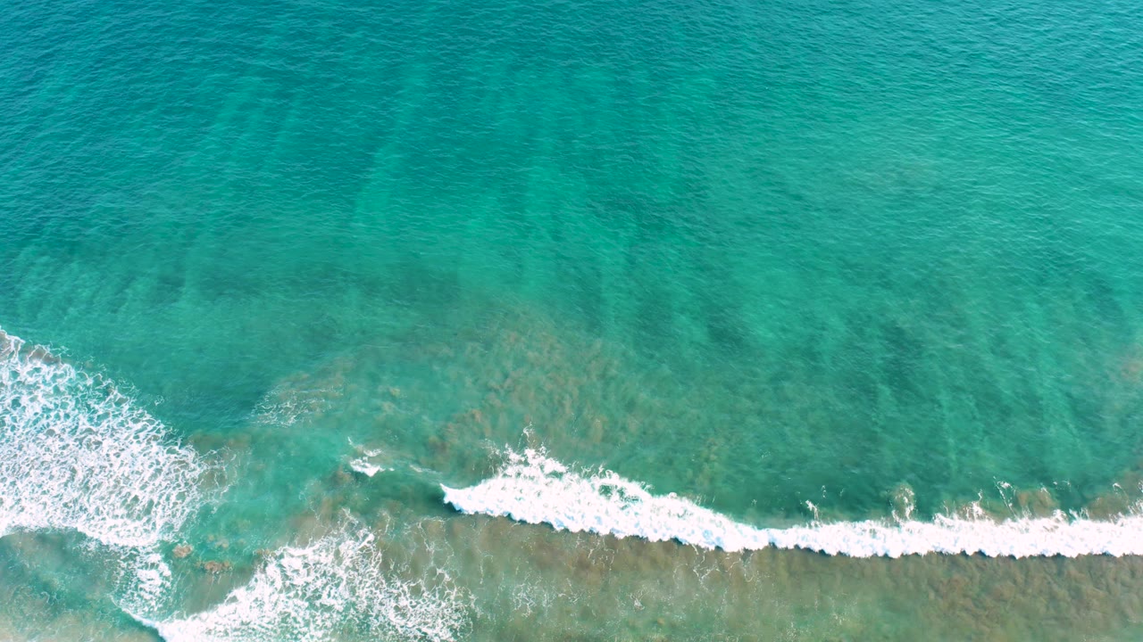 Top-down San Jose del Cabo ocean frame with a broad white foam line moving across blue-green water and darker underwater patches, with no visible people.
