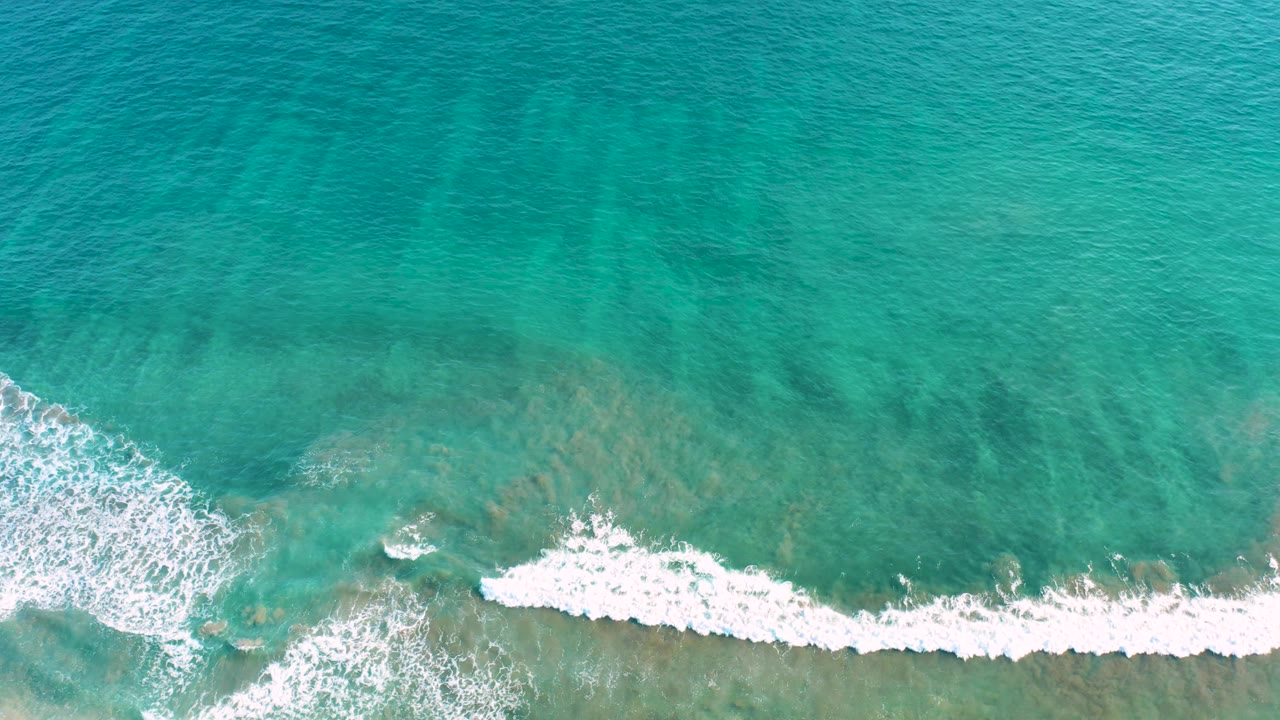 Aerial overhead frame from San Jose del Cabo showing angled white surf over turquoise water with tan-green seabed color visible below and no visible people.