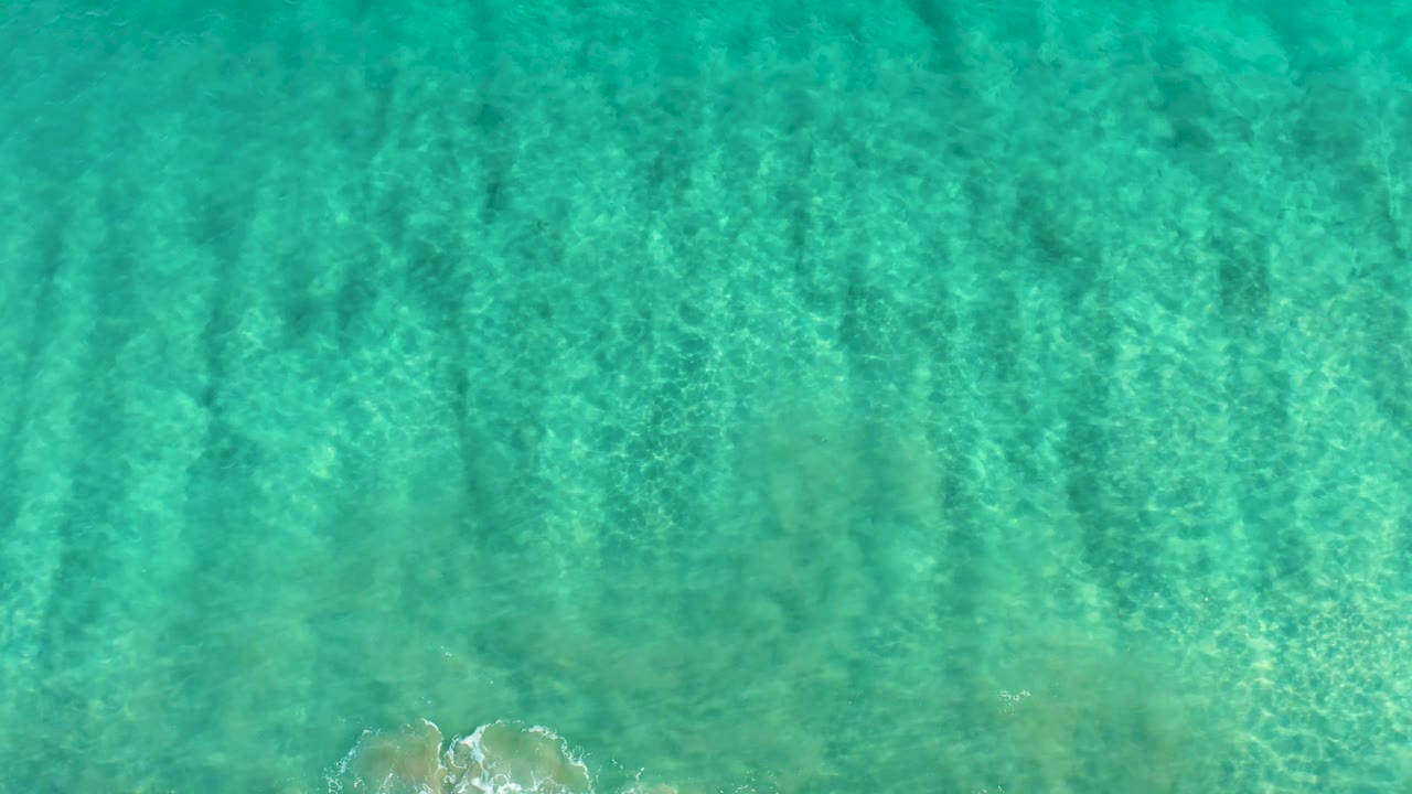 Overhead San Jose Del Cabo ocean water shows blue-green ripples, darker underwater streaks, and a pale rock patch near the lower edge with no visible people.