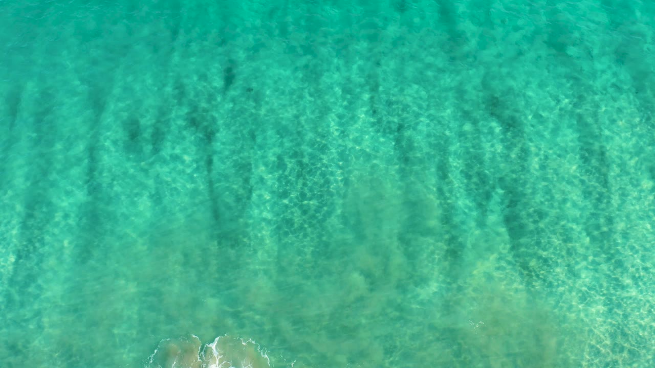 Top-down water view in San Jose Del Cabo shows turquoise surface ripples, mottled seabed detail, and a small pale patch along the lower edge with no visible people.
