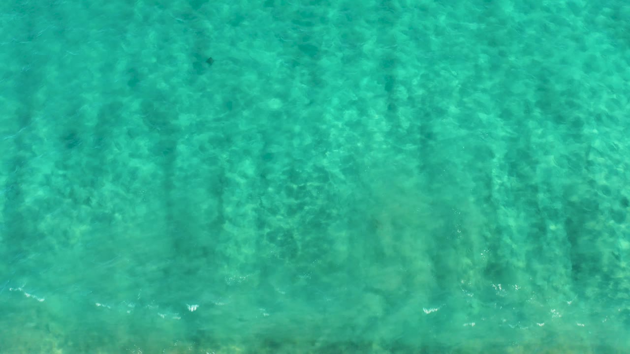 Top-down San Jose del Cabo water view with turquoise surface ripples, faint sandy bottom detail, small fish shapes, and no visible people.