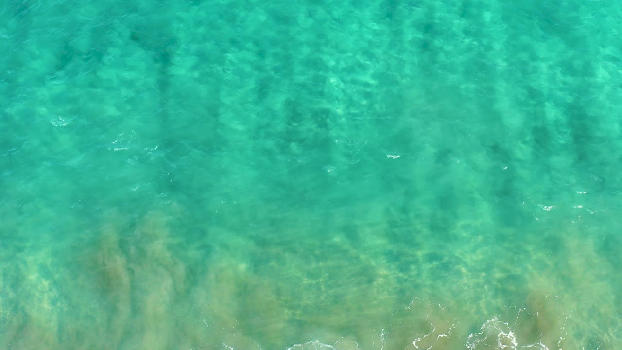 Transparent turquoise ocean water in San Jose Del Cabo reveals pale sand and darker underwater patches beneath a lightly rippled surface, with no visible people.