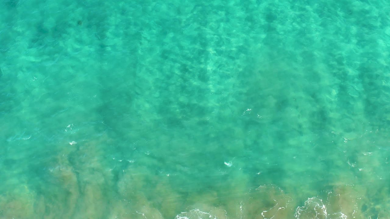 Overhead frame of shallow San Jose Del Cabo ocean water with pale sand visible beneath green-blue ripples and no visible people or structures.