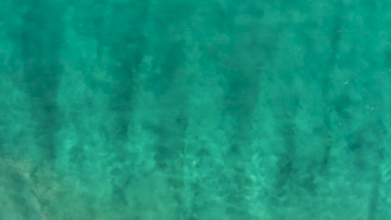 Clear blue-green ocean water in San Jose Del Cabo shows darker vertical shadow streaks falling across the pale sandy bottom, with no visible people.
