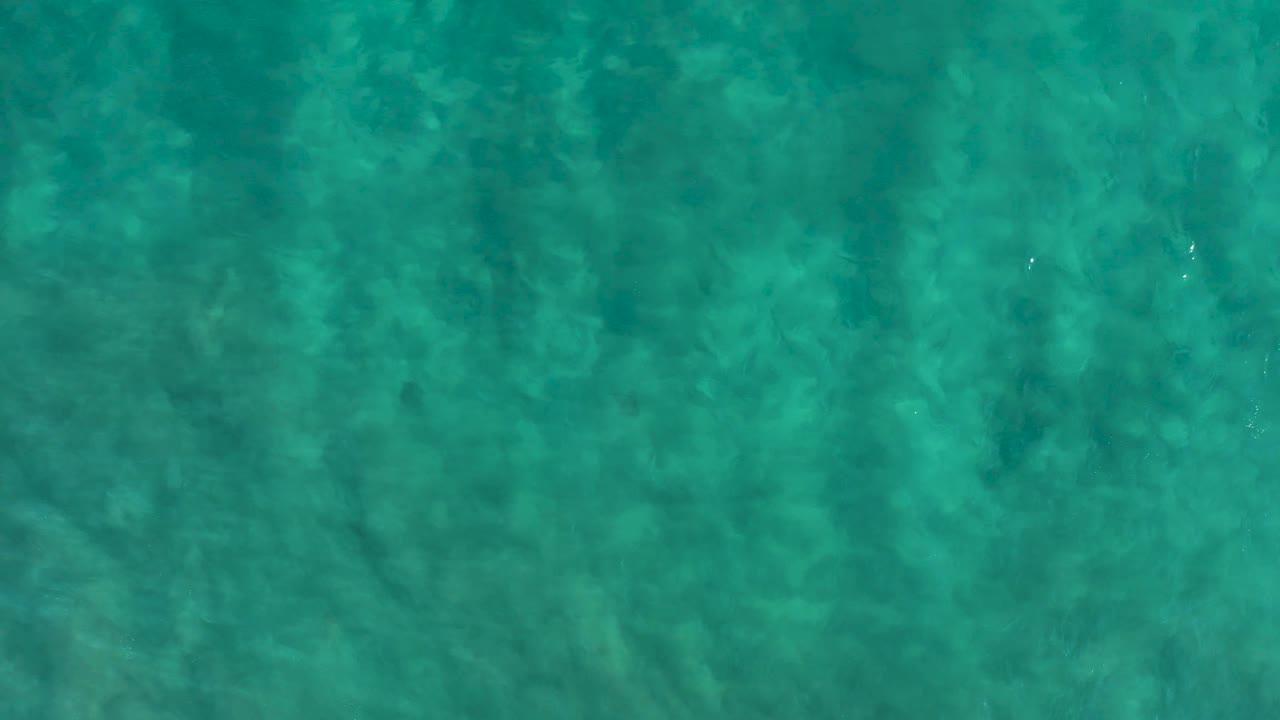 A straight-down view of shallow turquoise sea in San Jose Del Cabo shows faint reef-like floor patterns under clear water and no visible people.