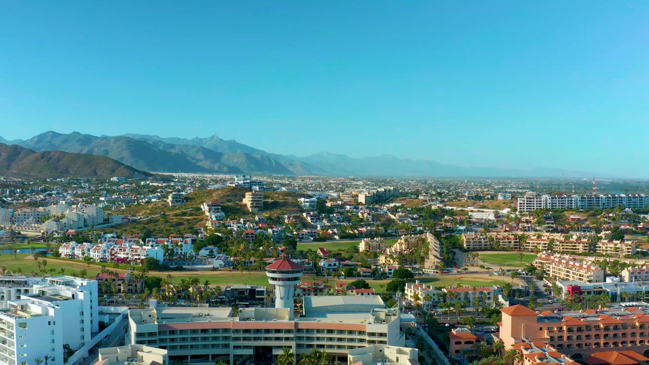 Opening frame of San Jose del Cabo, Baja California Sur, with white buildings, green fields, distant mountains, clear blue sky, and no visible people.