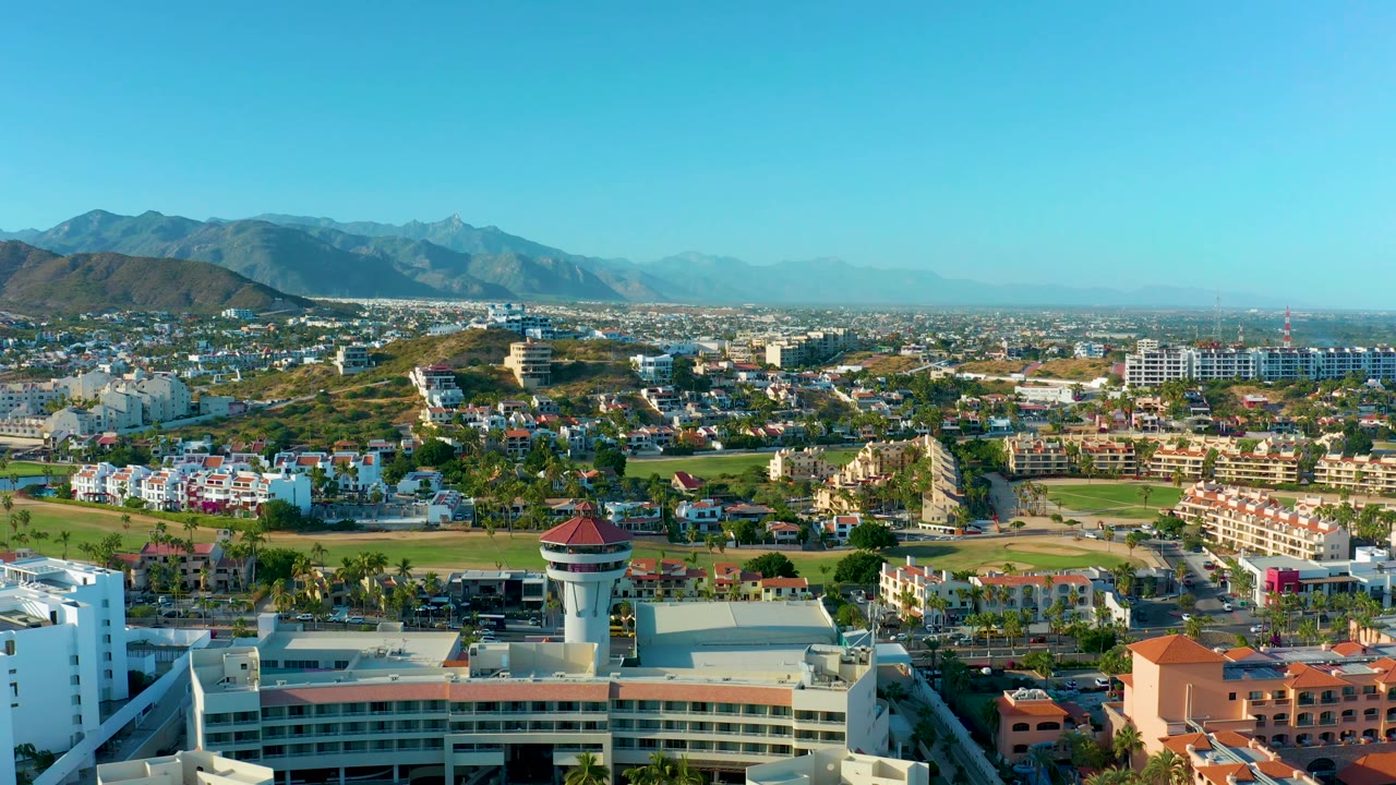 Aerial frame in San Jose del Cabo with foreground rooftops, a central red-roof tower, dense residential and commercial blocks, green space, hazy mountain ridges, and no visible people.