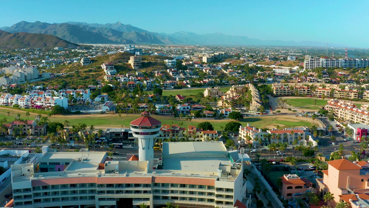 A wide aerial view of San Jose Del Cabo shows a round-topped water tower above nearby roofs, low-rise neighborhoods, green fields, distant mountains, clear blue sky, and no visible people.