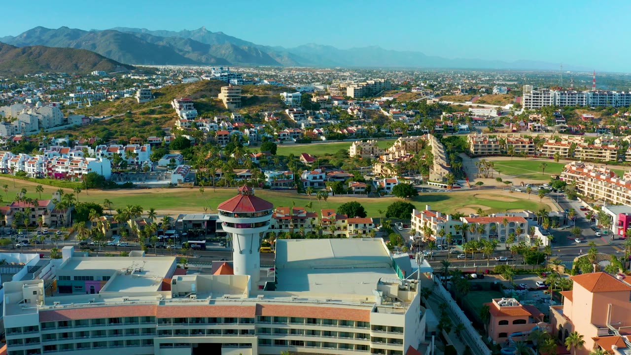 The San Jose Del Cabo aerial view shows the water tower near the lower frame, a green field and clustered buildings in the midground, mountains along the horizon, and no visible people.