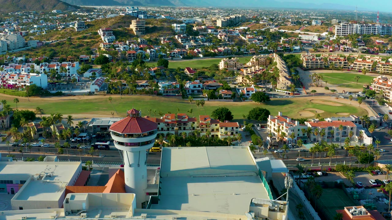 Aerial frame over San Jose del Cabo showing a red-roofed control tower in the foreground, a green open field beyond it, residential blocks, roads with cars, hills in the distance, and no visible people.