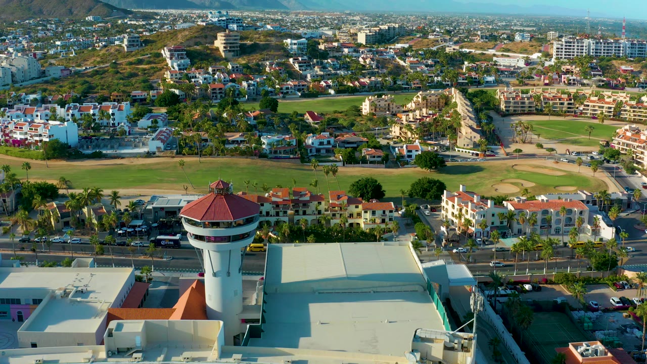 San Jose del Cabo aerial view with the control tower slightly right of center, pale rooftops in front, a green field and sand areas behind, roadside vehicles, residential buildings, and no visible people.