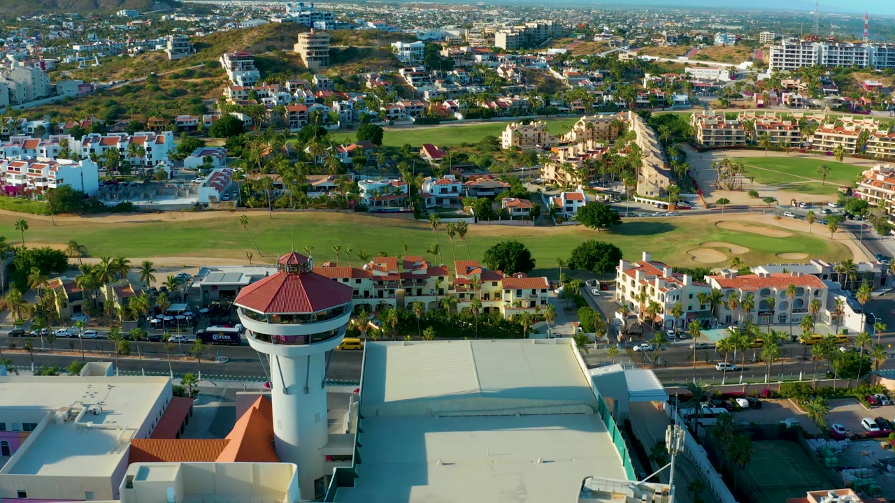 Aerial frame in San Jose del Cabo with the control tower near the lower center, a wide green park-like field across the middle, roads and cars below, dense residential buildings, hills, and no visible people.