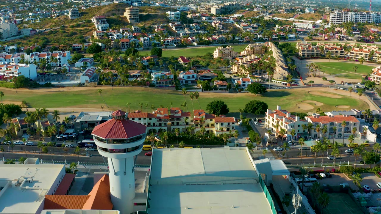 Aerial view with a red-roofed tower beside a large roof, green field, residences, and road traffic.