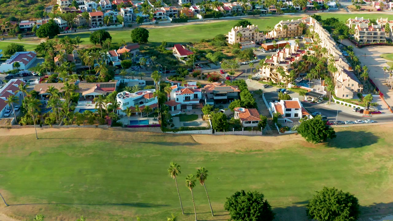 Opening aerial frame over a San Jose Del Cabo residential community, with a wide green golf course foreground, palm trees, villas, and no visible people.