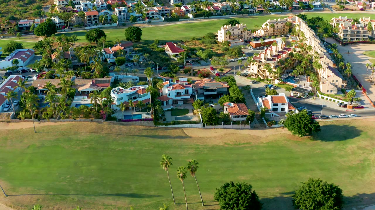 San Jose Del Cabo aerial frame with fairway grass across the lower half, a palm-lined residential street, clustered homes, parked cars, and no visible people.