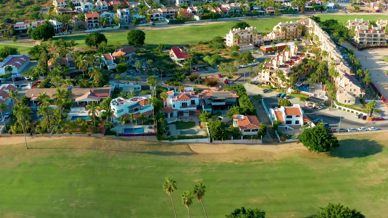 Aerial frame in San Jose Del Cabo showing a broad golf course foreground, rows of homes beyond the road, palms, landscaped yards, and no visible people.